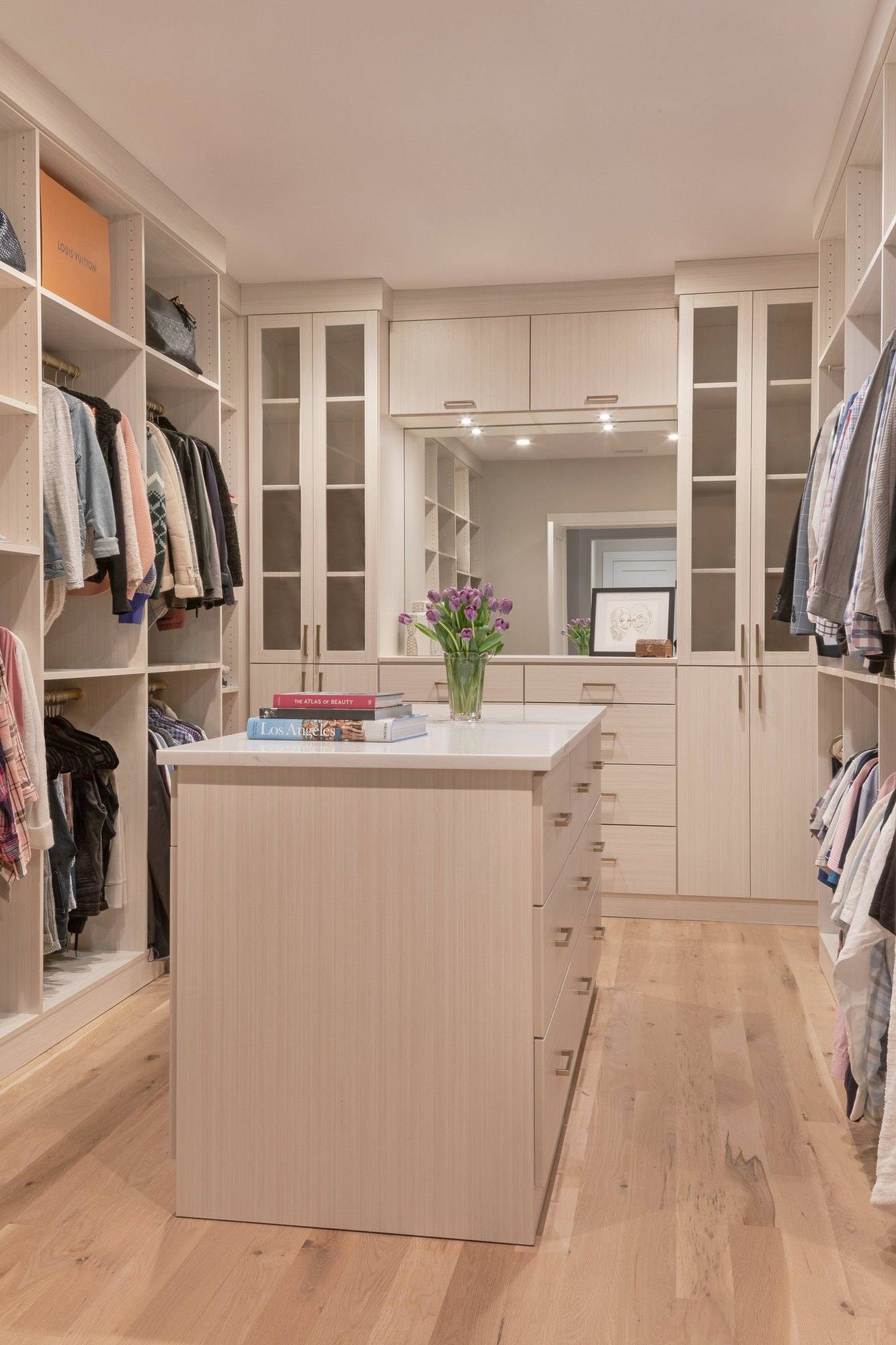 Long walk-in closet in a pale wood-grain finish with a center island, glass-front cabinets and lighted shelving