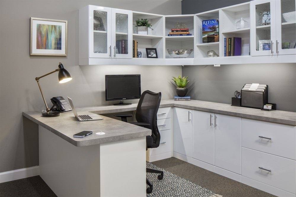 White home office with an L-shaped desk, lower drawers and file storage, and glass-front upper cabinets