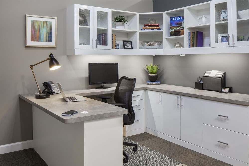 White home office with an L-shaped desk, lower drawers and file storage, and glass-front upper cabinets