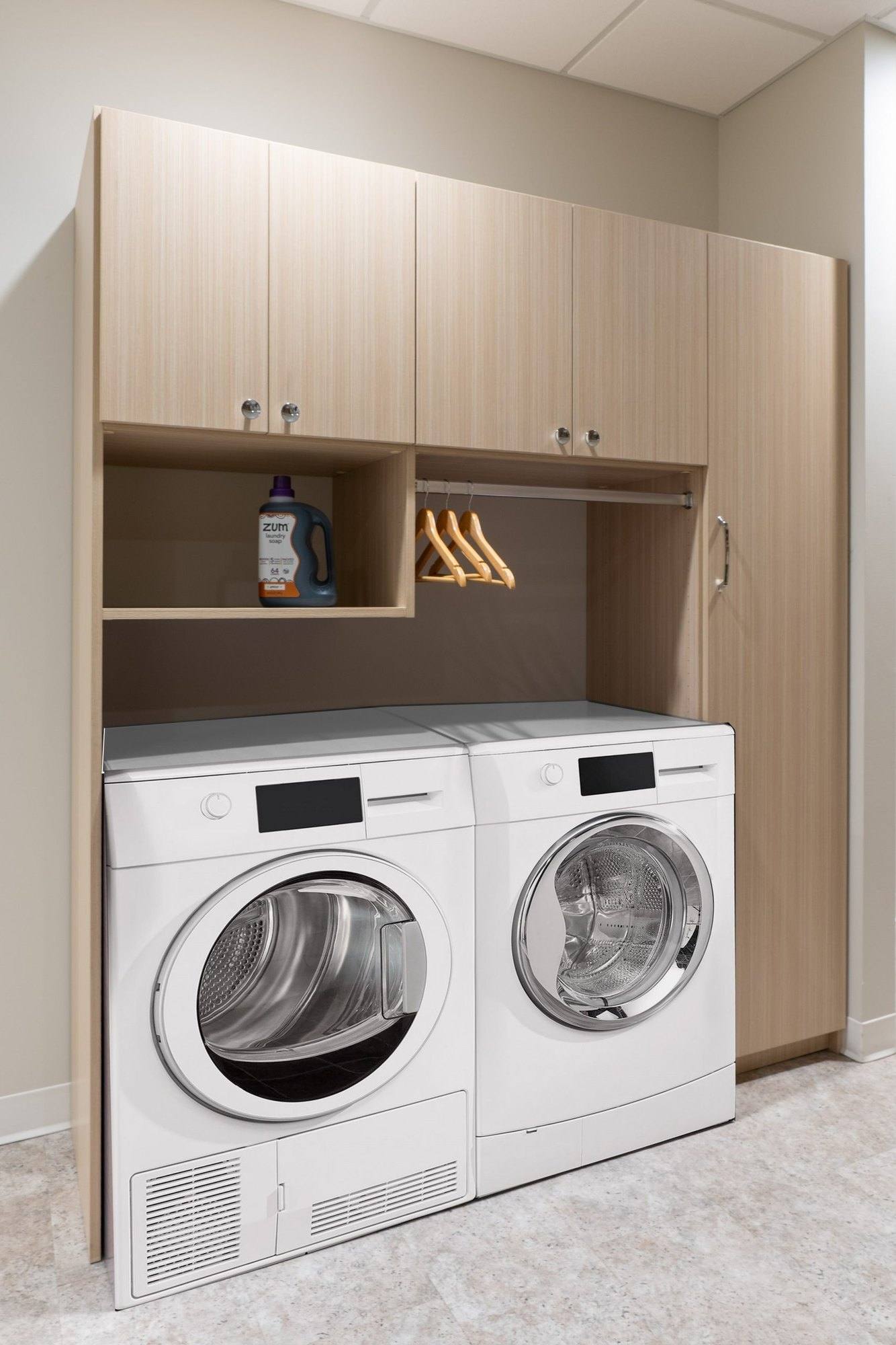 Laundry display in the showroom with wood-tone cabinets above a washer and dryer, an open cubby and a short hanging rod